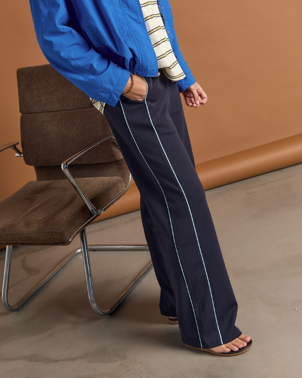 Person wearing a blue jacket and black pants with blue stripes, standing in front of a brown chair.